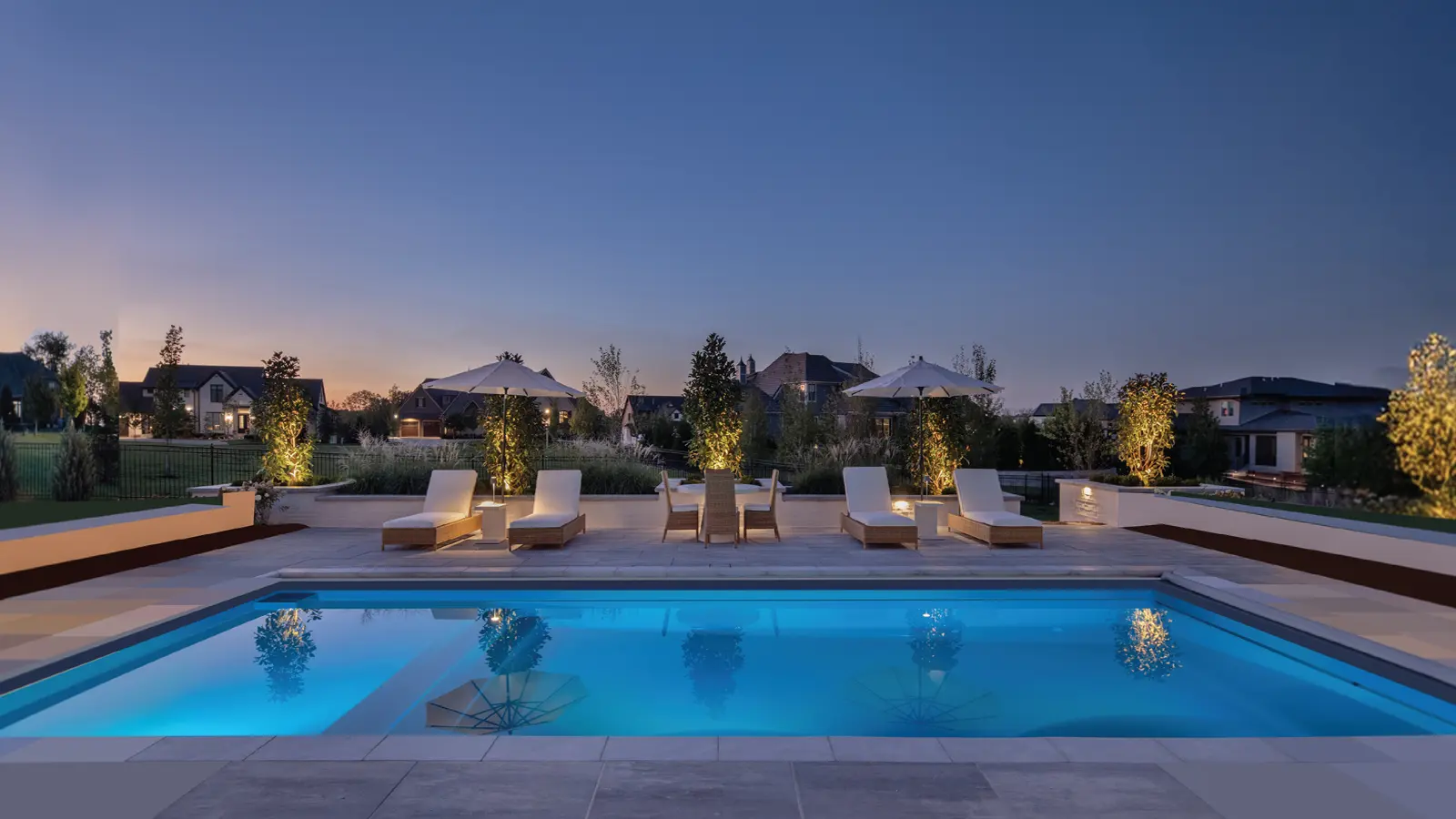 AvivaPools_inset01_2026 Aviva Pools luxury fiberglass pool and spa at dusk, illuminated with blue underwater lighting and flanked by white lounge chairs and lush landscaping