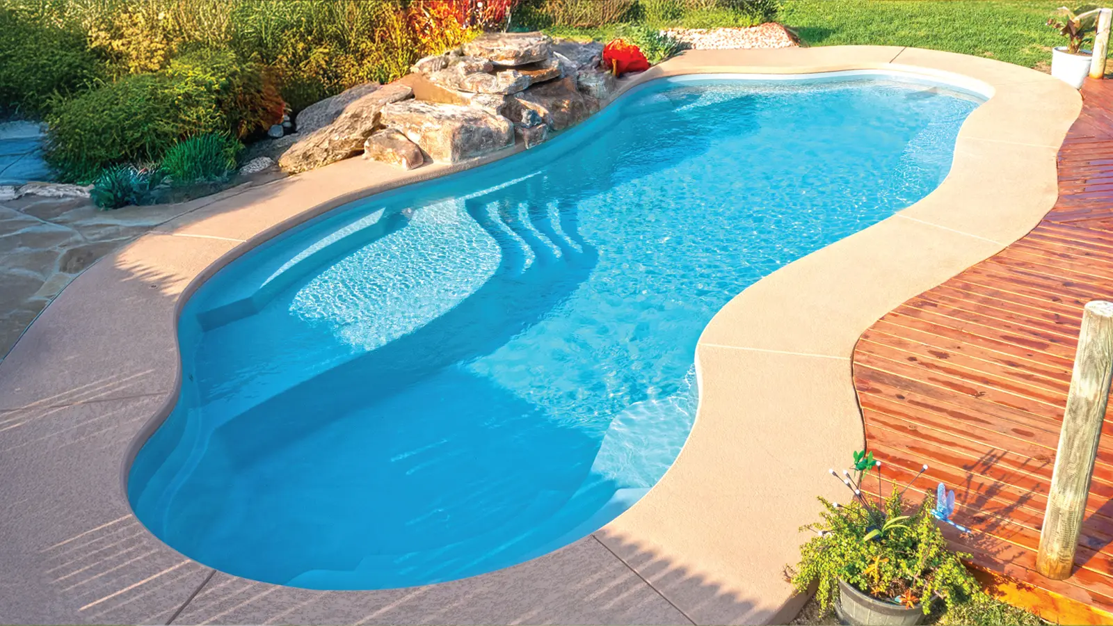 AvivaPools_inset04_2026 Aviva Pools freeform fiberglass pool with natural rock waterfall feature and wood deck, nestled in a lush backyard landscape
