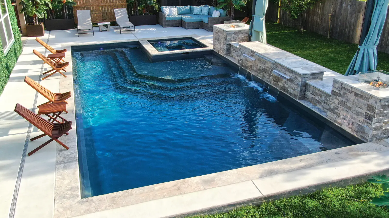 AvivaPools_inset05_2026 Aviva Pools rectangular fiberglass pool with attached spa and cascading stone waterfall feature, surrounded by wooden lounge chairs and a private backyard setting