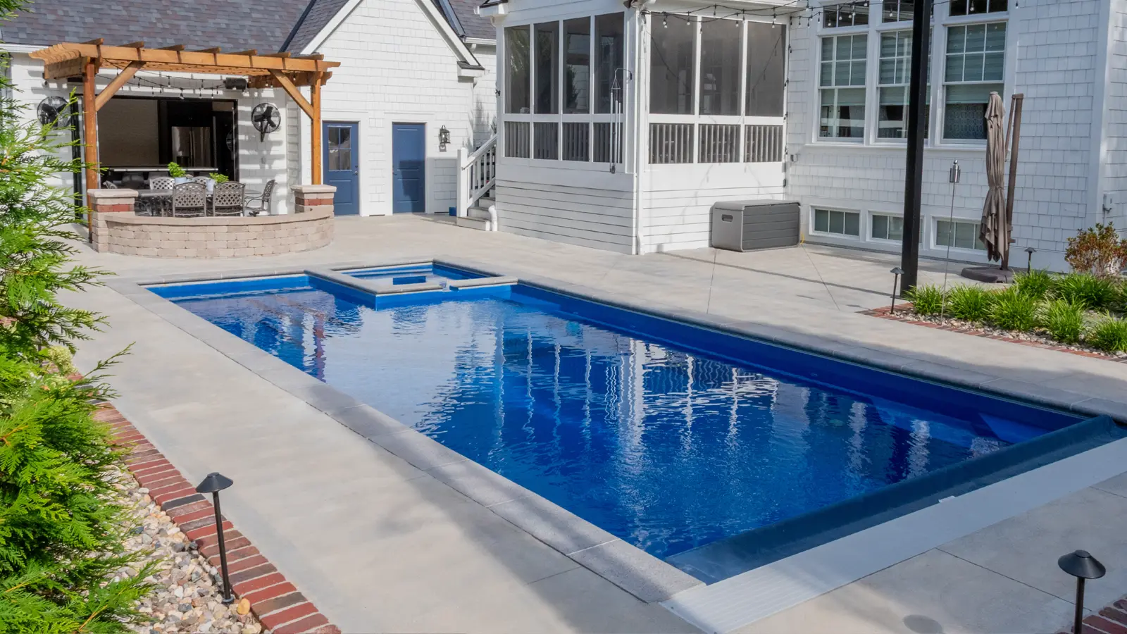 AvivaPools_inset07_2026 Aviva Pools rectangular fiberglass pool with attached spa in deep navy, installed in the backyard of a white craftsman-style home with mature landscaping