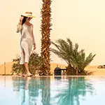 Woman in a white dress walking beside an Evo Pools resort-style pool at golden hour, with palm trees and desert dunes stretching into the distance